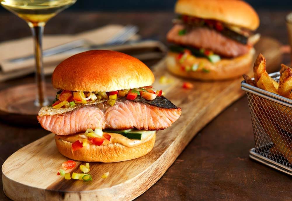 A photo of a salmon fillet inside a burger bun on top of a wooden board