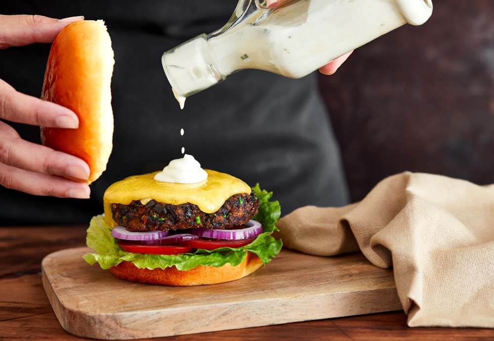 A hand dripping sauce on top of a burger, on a wooden serving board with a tea towel next to it Ideal For Valentine's Day Quiz