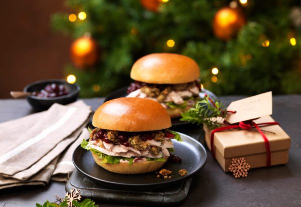 Two turkey, stuffing and cranberry burgers on plates in front of a Christmas tree with a Christmas present next to them Christmas Food Trends