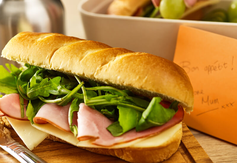 A close up of a soft brioche baguette filled with lettuce, cheese and ham also called Jambon Fromage in frenchnext to an orange post-it note and lunch box